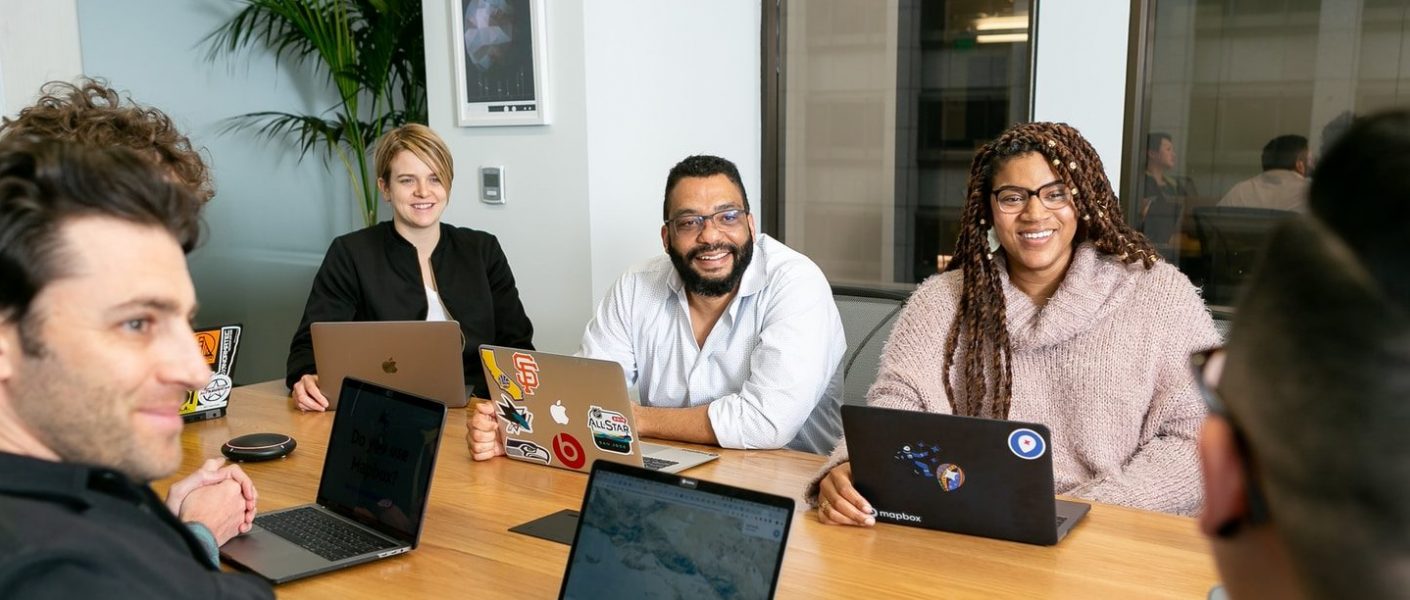 four people all on laptops, two men and two women, listen to person talking in a board meeting
